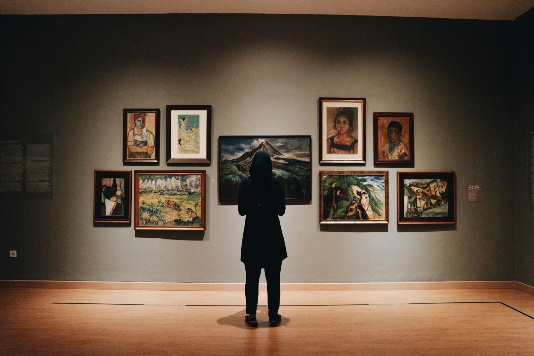 A woman standing alone in a gallery, looking at a wall of paintings.