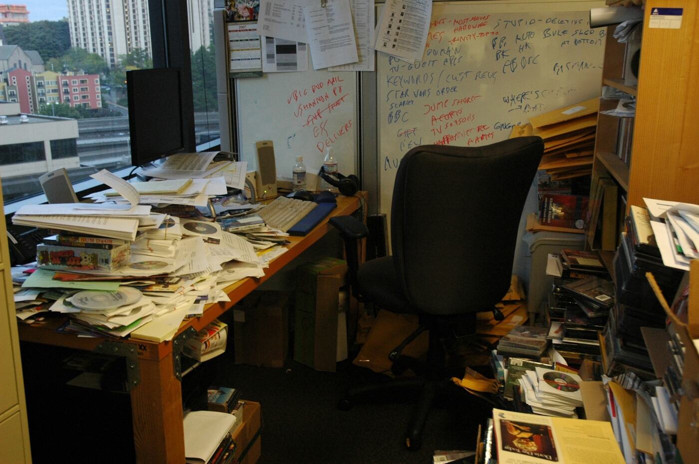 A cluttered office desk by a city window: papers, notes, a whiteboard of handwriting.
